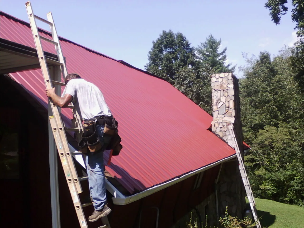 Roofer on a ladder beside a red metal roof and stone chimney — Soffit & Fascia Repair by Roofing Brothers in Allston